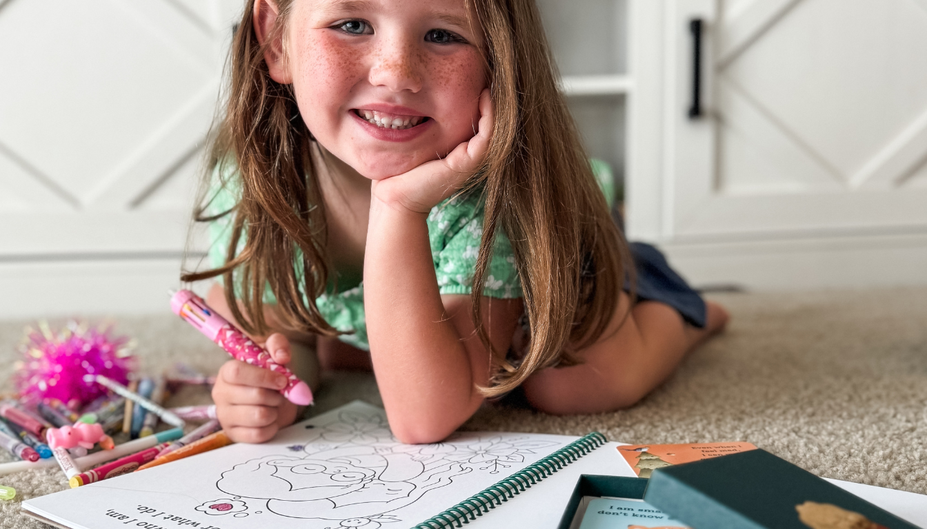 Young girl lying on the floor with coloring books and crayons, smiling.