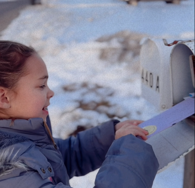 Person in a blue coat opening a mailbox outdoors with snow on the ground.